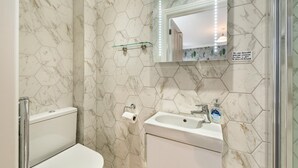 A bathroom with hexagonal marble tiles, a glass shelf, a toilet, a sink with a soap dispenser, and a shower with a sign.
