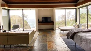 A modern bedroom with a large bed, a seating area, a TV, and a view of the ocean.