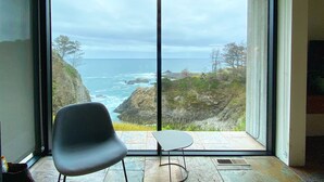A room with a large sliding glass door leading to a balcony with a view of the ocean and rocky coastline.