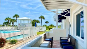 A poolside area with a covered seating area, a pool, and palm trees in the background. - Elite 1%~Private Patio~Pool View~Oceanfront~Hot Tub~Beach Gear! (Cape Canaveral)