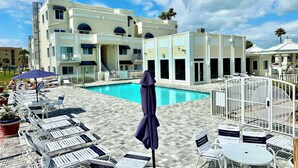 A poolside area with lounge chairs, a swimming pool, and a multi-story building with balconies. - Elite 1%~Private Patio~Pool View~Oceanfront~Hot Tub~Beach Gear! (Cape Canaveral)