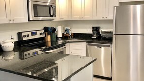 A modern kitchen with white cabinets, stainless steel appliances, a granite countertop, and a central island with stools. - Elite 1%~Private Patio~Pool View~Oceanfront~Hot Tub~Beach Gear! (Cape Canaveral)