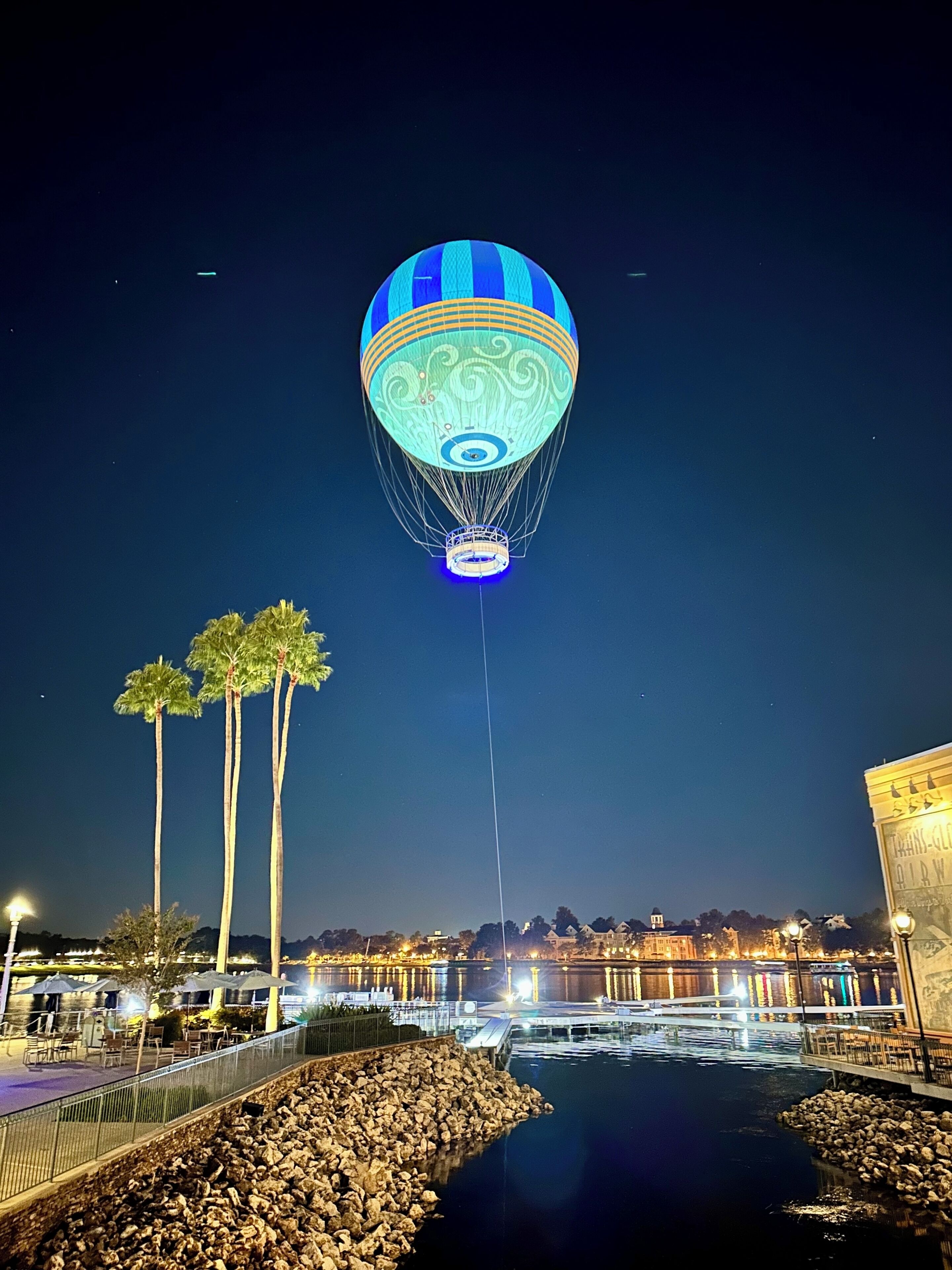 A hot air balloon with a blue and white design, tethered to a structure, over a waterfront area with palm trees and buildings in the background.
