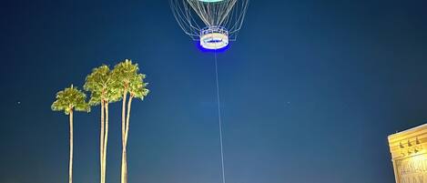A hot air balloon with a blue and white design, tethered to a structure, over a waterfront area with palm trees and buildings in the background.