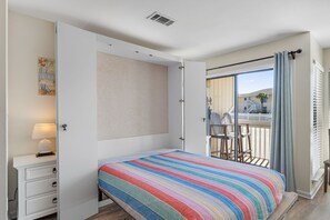 Une chambre à coucher comprenant un lit, une table de chevet, une commode, une fenêtre donnant sur un balcon et une bouche d’aération au plafond.