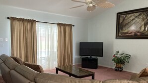 A living room with a leather sofa, a flat-screen TV, a ceiling fan, and a large painting on the wall. - Beautiful Home With Heated Pool In A Nice Neighborhood (Spring Hill)