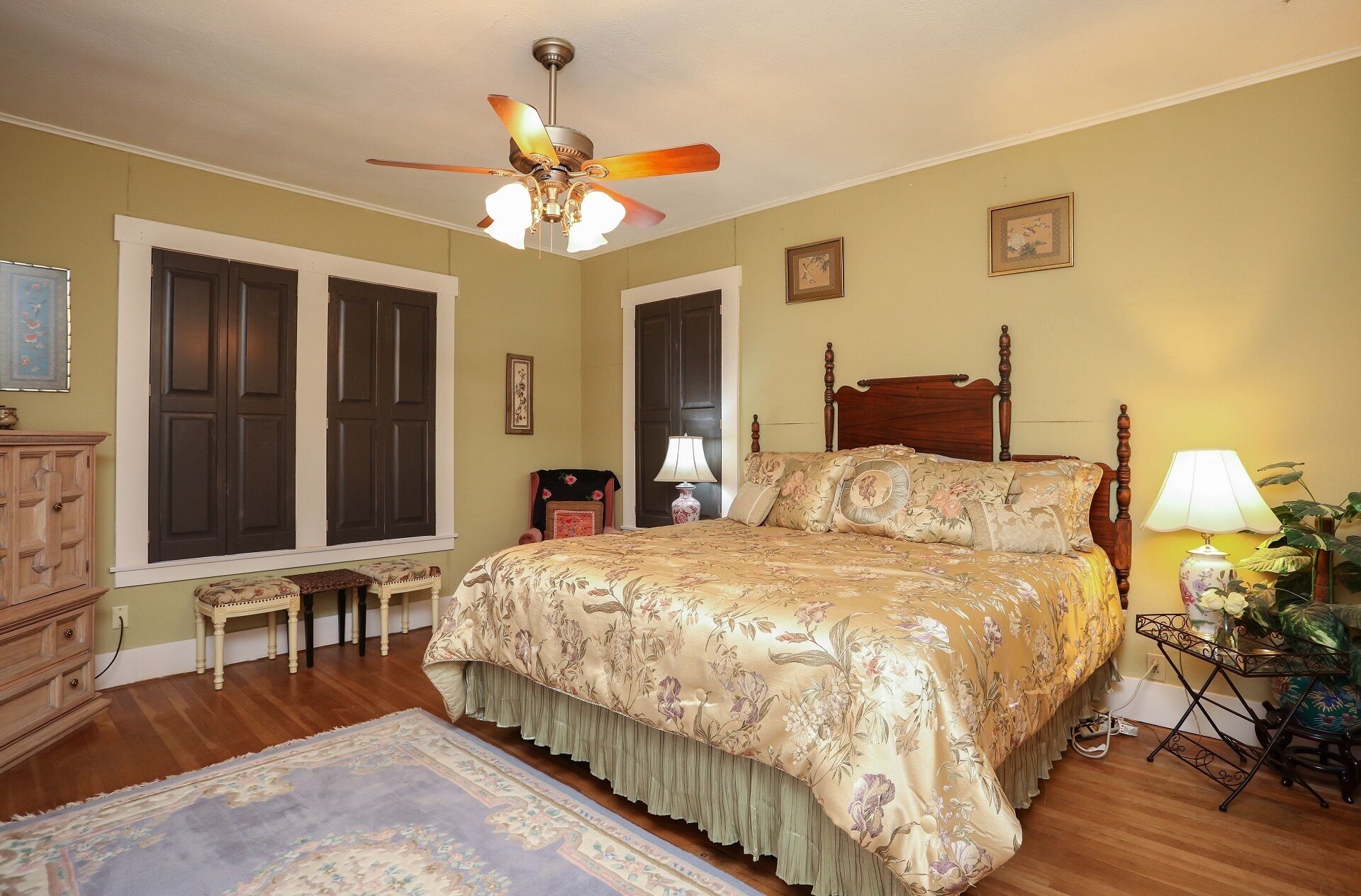 A bedroom with a large bed, a ceiling fan, a nightstand, a lamp, a dresser, and a window with shutters.