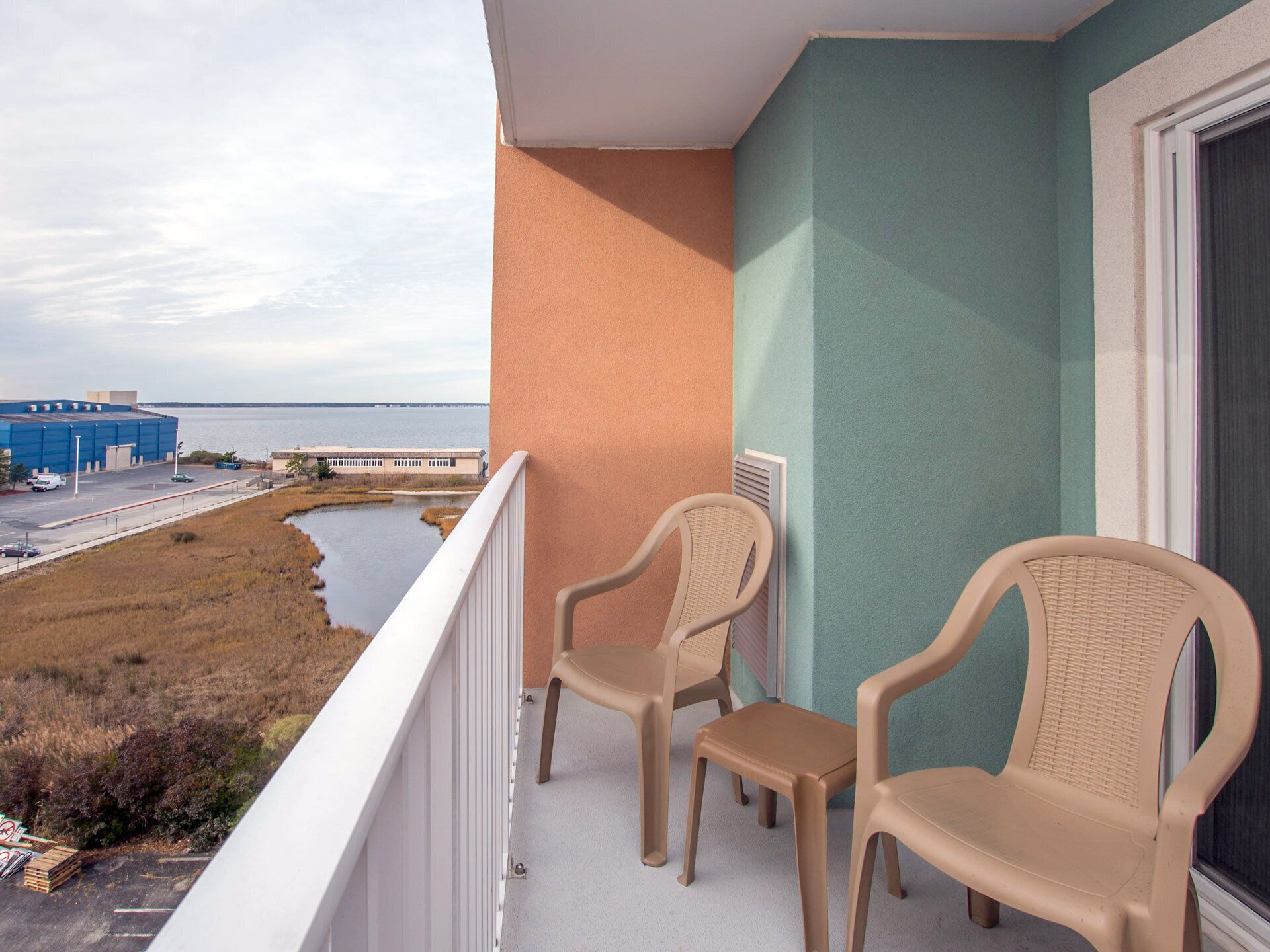 A balcony with two wicker chairs and a small table, overlooking a body of water and industrial buildings.