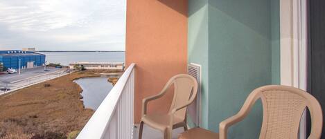 A balcony with two wicker chairs and a small table, overlooking a body of water and industrial buildings.