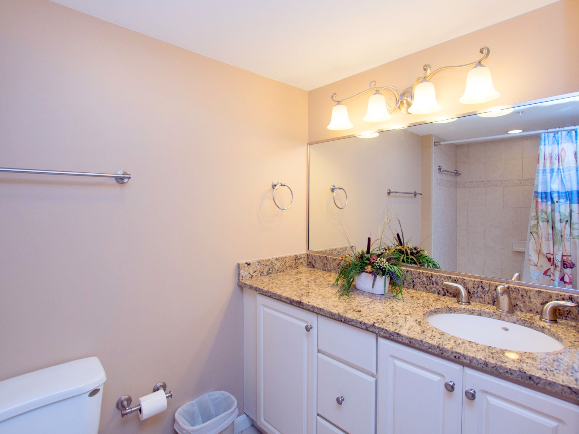 A bathroom with a granite countertop sink, a mirror with three lights, a towel rack, and a toilet.