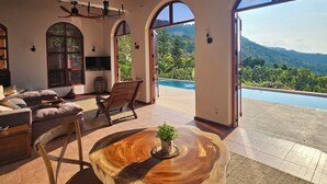Ein großzügiger Wohnbereich mit einem großen Fenster, das einen Pool und Berge blickt, Holzmöbel und einem Deckenventilator.