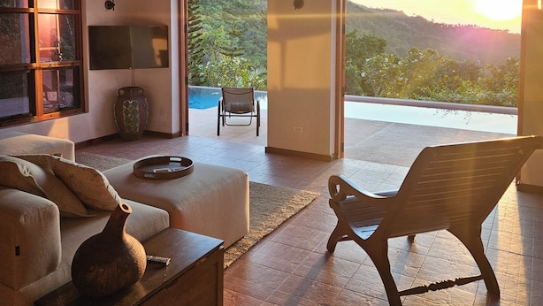 Ein geräumiges Wohnzimmer mit einer Couch, einem Couchtisch und einem Sessel. Ein Fernseher steht auf einem Schrank, eine dekorative Vase ist vorhanden, und ein großes Fenster bietet Blick auf eine üppige Landschaft.
