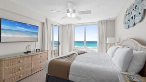 Habitación de hotel con cama, televisor, ventilador de techo, vista a la playa y un adorno en la pared.