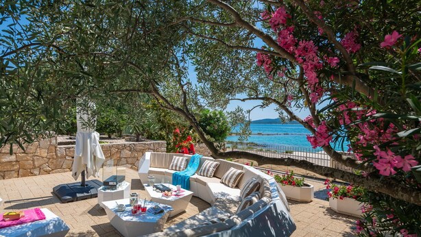 A sunlit outdoor seating area with a curved sofa, tables, and chairs, overlooking a coastal view.