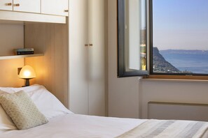 A bedroom with a bed, a nightstand with a lamp, a window with a view of the sea, and a shelf with books.