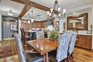 A spacious dining area with a wooden table and patterned chairs, a chandelier, and wooden beams on the ceiling.