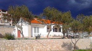 A white building with a red-tiled roof, stone walls, and trees in front.