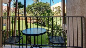 A balcony with a round table and two chairs, overlooking a well-maintained garden with palm trees and a clear blue sky.