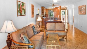 A spacious living room with a sofa, chairs, and a coffee table. There is a ceiling fan, a kitchen area with wooden cabinets, and a dining table.