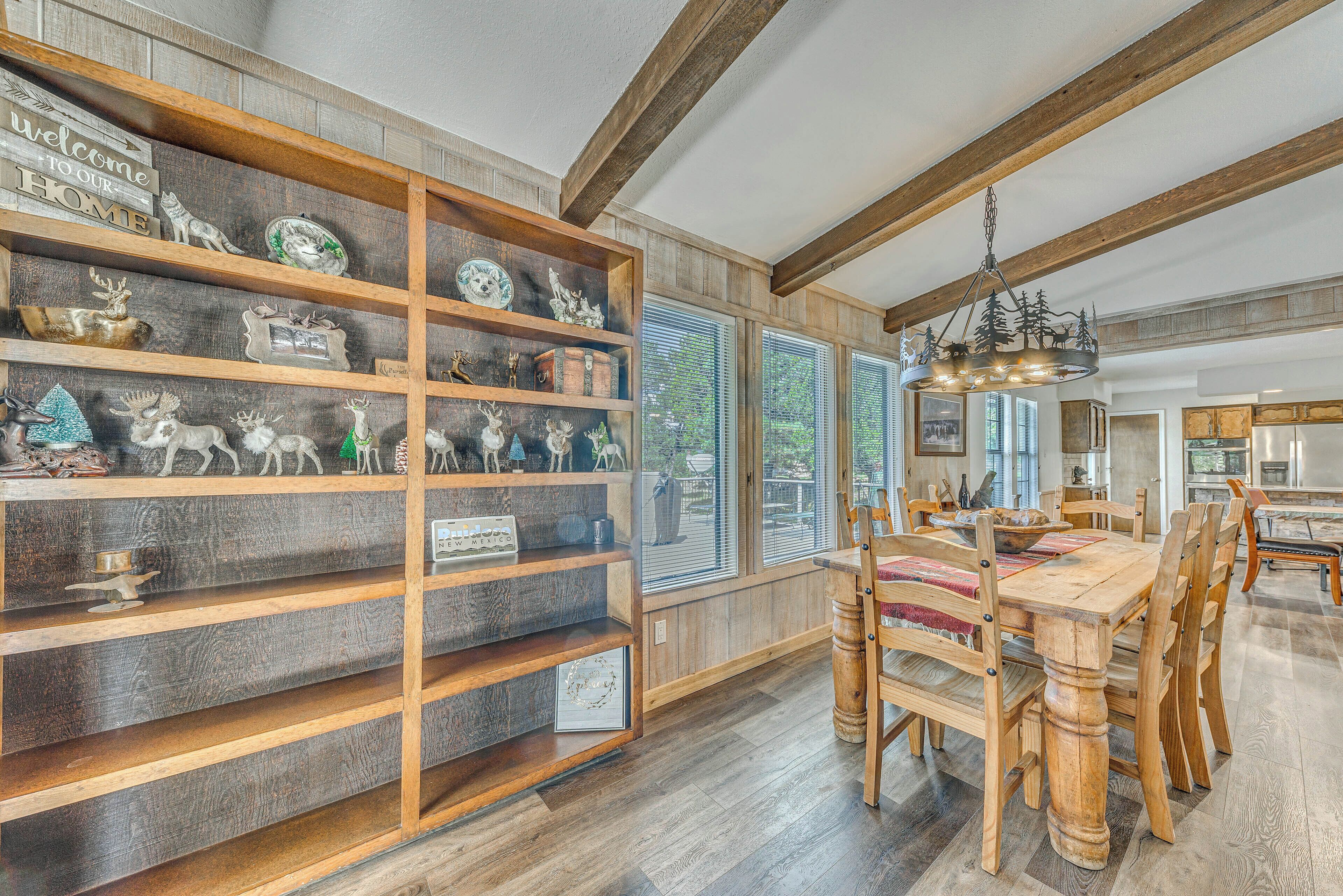 A dining area with a wooden table and chairs, a wooden bookshelf with various items, and a view of the outdoors through large windows.