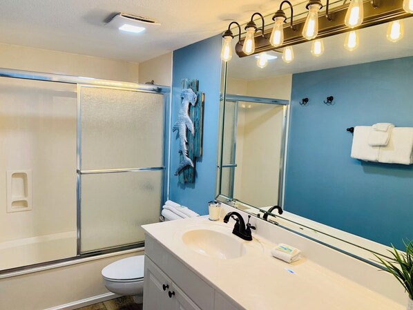A bathroom with a glass shower enclosure, a white sink with a black faucet, a large mirror with lights, and a towel rack with towels. - Beachside 2BR | Free Beach Service & Sunset Views (Destin)