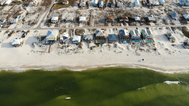 An aerial view of a coastal resort with multiple beachside cottages, a main building with a pool, and a road running parallel to the beach. - Reserve now. Beautiful pool and ocean. Easy social distancing. Huge pool deck. <br> (Fort Morgan)