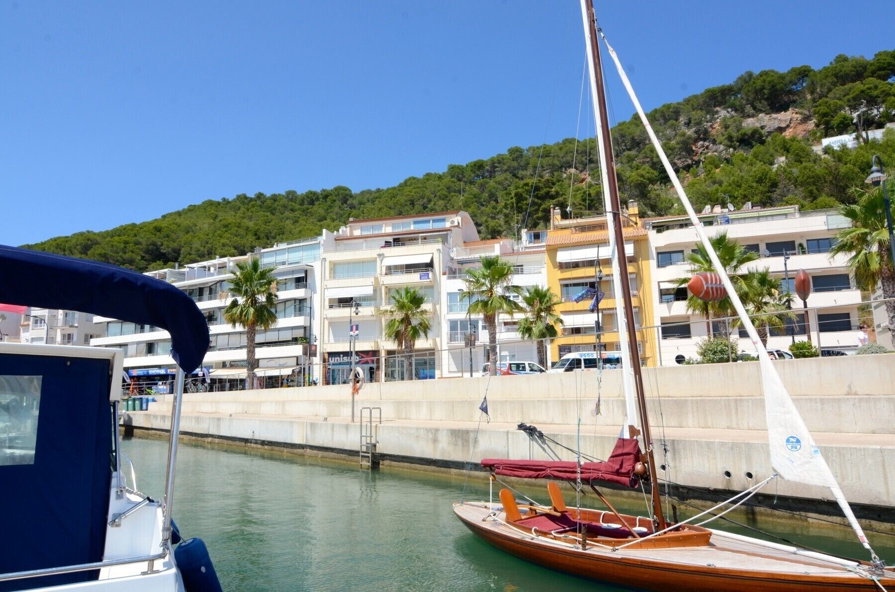 Un port esportiu amb embarcacions amarrades, un moll de formigó i edificis al fons.