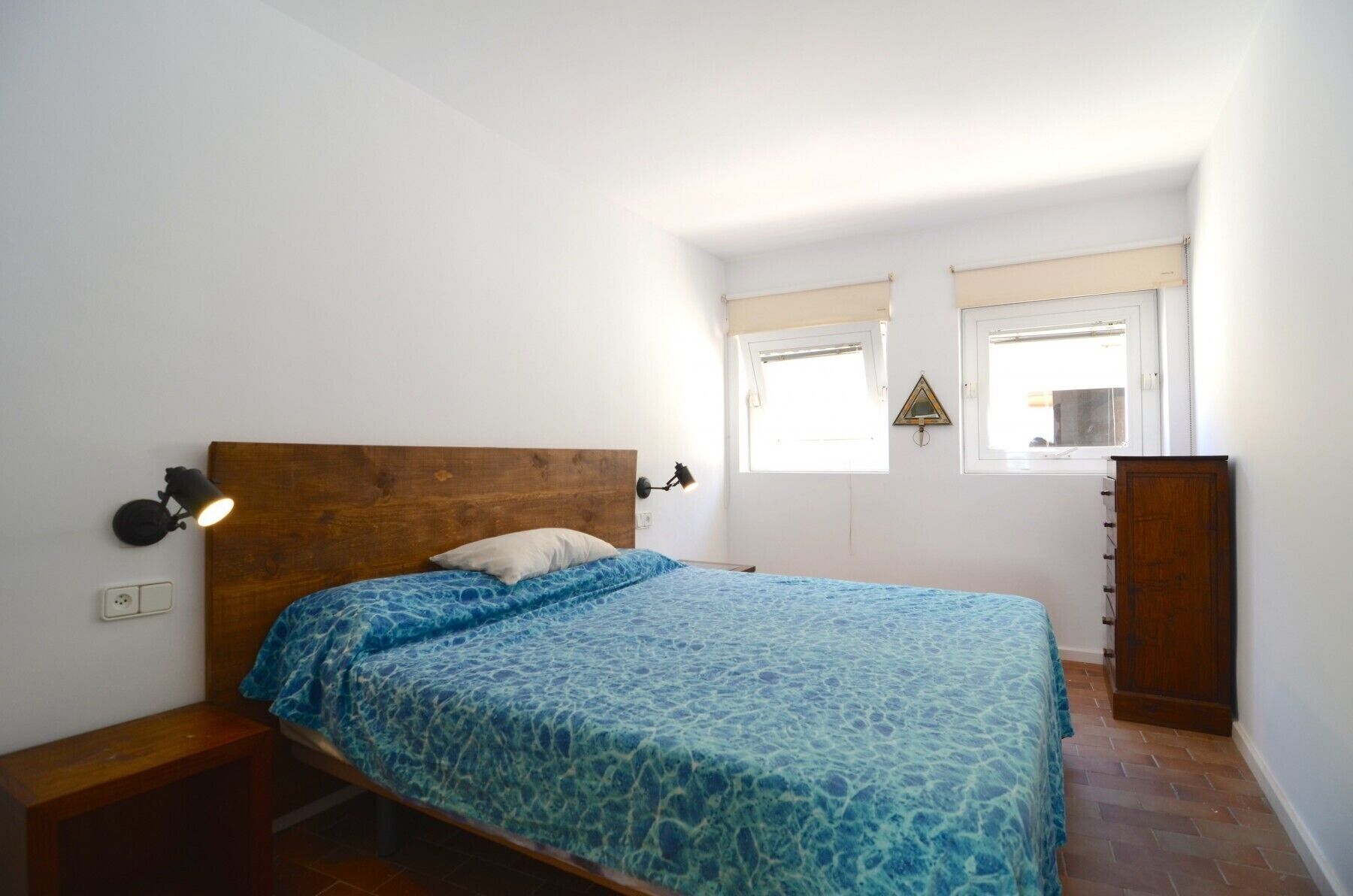 A bedroom with a wooden headboard, a bed with a blue patterned bedspread, two wall-mounted lamps, a wooden dresser, and a window with a view.