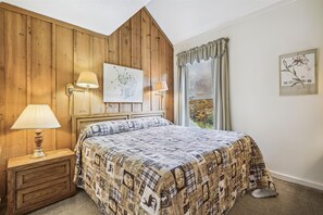 Een slaapkamer met een houten hoofdeinde, een bed met een beddengoed met een patroon, twee wandlampen, een ingelijst schilderij boven het bed en een raam met gordijnen.