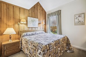 A bedroom with a wooden headboard, a bed with a patterned bedspread, two wall-mounted lamps, a framed picture above the bed, and a window with curtains.