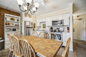 Een moderne keuken met witte kastjes, een roestvrijstalen koelkast, een magnetron en een fornuis. Er staat een houten eettafel met zes stoelen.