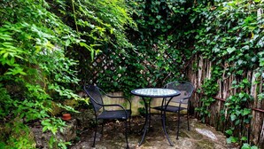 A small outdoor seating area with a round table and two chairs, surrounded by dense greenery and a wooden fence.