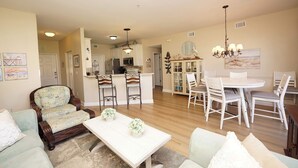 A spacious living room with a large sofa, a dining area with a white table and chairs, and a kitchen in the background.