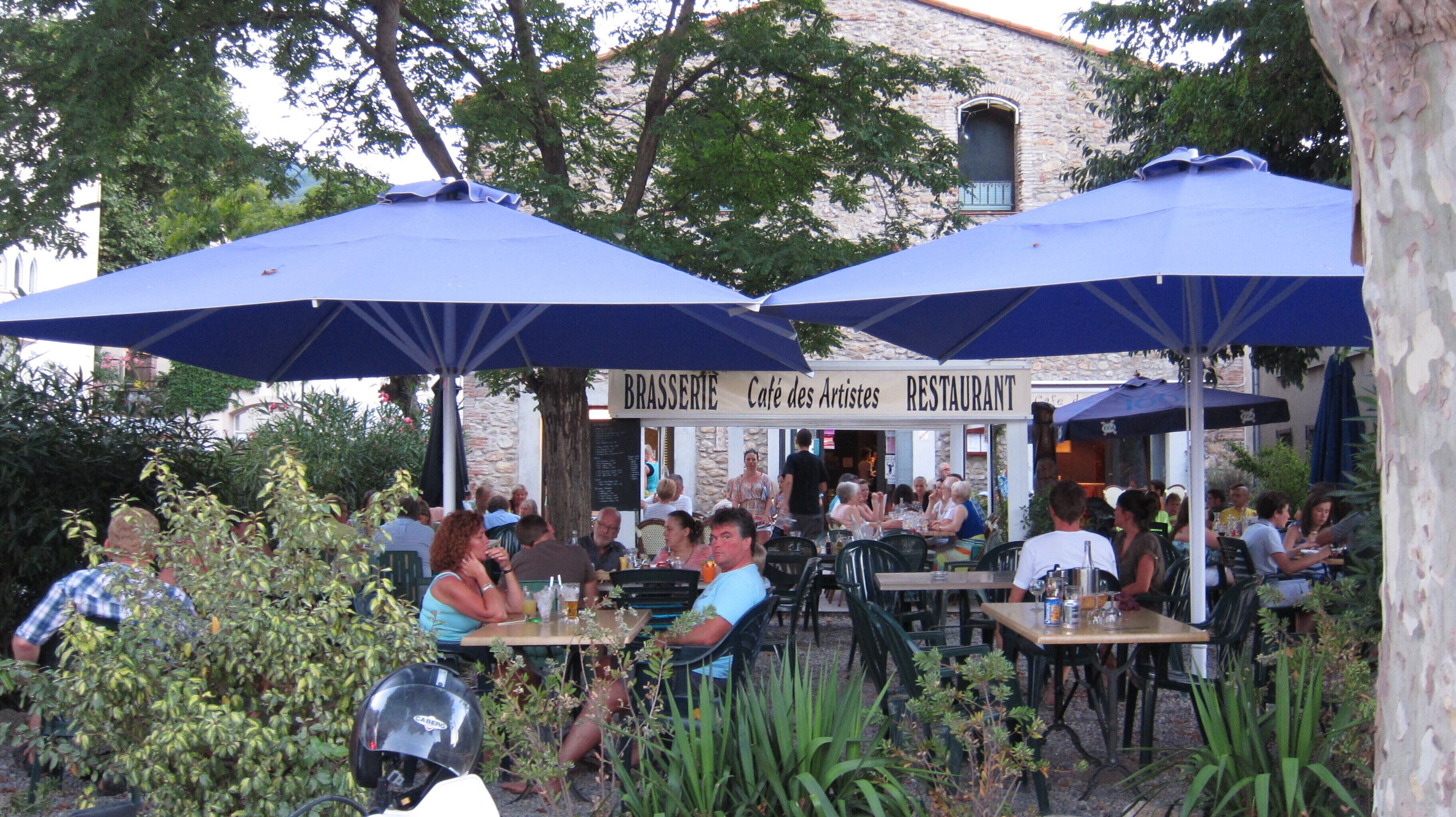 Un café en plein air avec des tables et des chaises sous des parasols bleus, entouré de verdure.