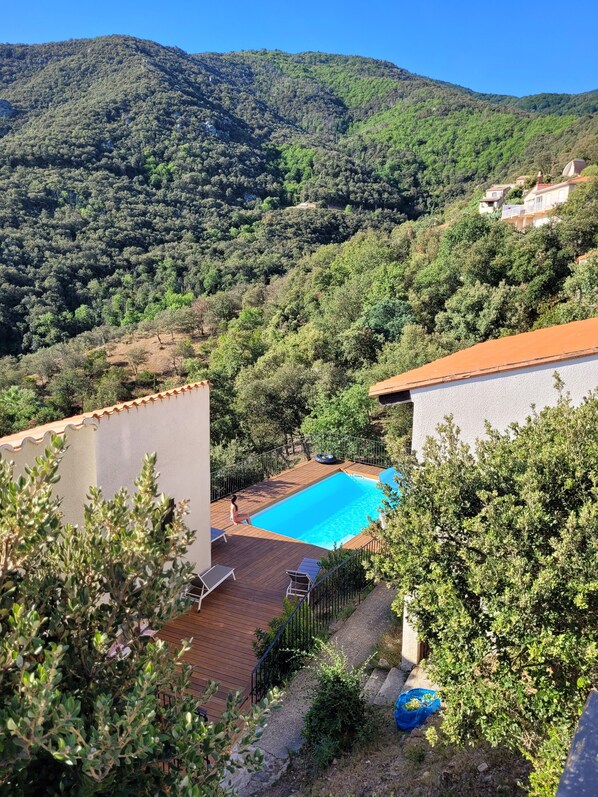 A pool area with a wooden deck, surrounded by greenery and a mountain backdrop.
