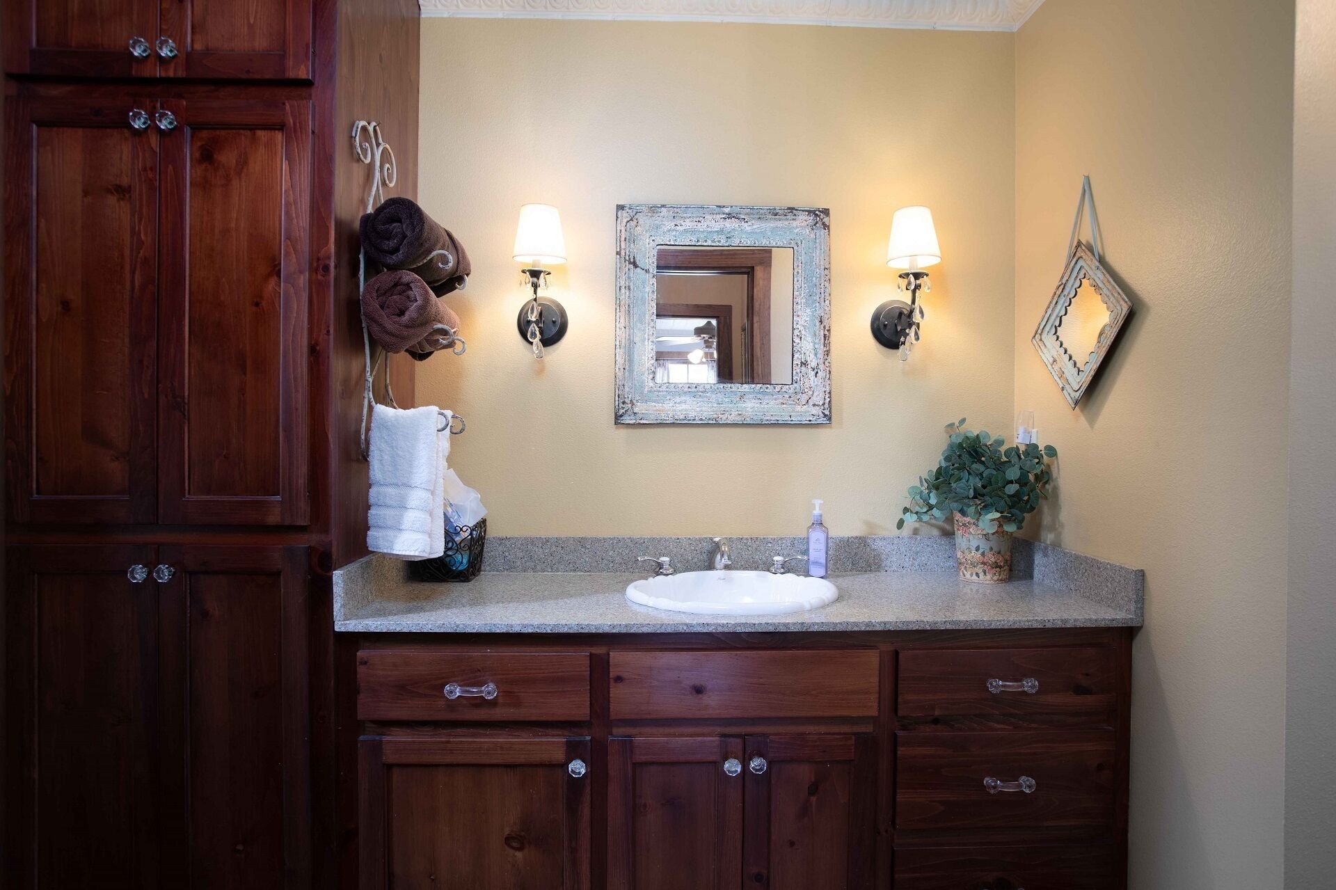 A bathroom with a wooden vanity, a mirror, two wall-mounted lights, a sink, and a potted plant.