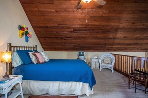 Une chambre avec un plafond en pente, un lit avec une literie bleue, une lampe de chevet, une petite table, une chaise, un téléviseur et un élément décoratif mural.