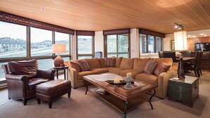 Ein geräumiges Wohnzimmer mit einem großen Ecksofa, Ledersesseln und einem Holztisch. Große Fenster bieten einen Blick ins Freie.
