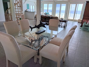 A dining area with a glass table surrounded by striped chairs, a decorative shark sculpture, and a view of the ocean through large windows. - Little Cayman 3 Bedroom Oceanfront  Private Villa (West End)
