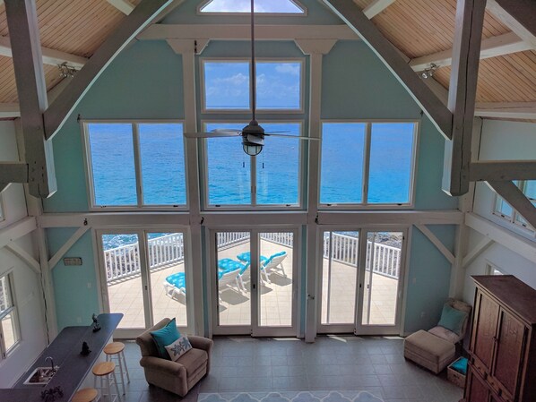 A spacious room with a high ceiling, large windows offering a view of the ocean, a ceiling fan, and a balcony with outdoor seating.