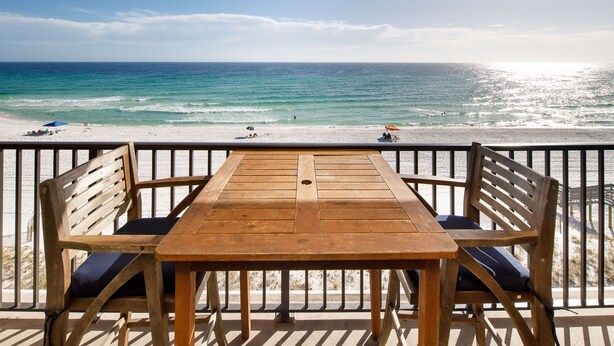 Ein Holztisch und -stühle auf einem Balkon mit Blick auf einen Strand und das klare Meer, sowie badende Personen.