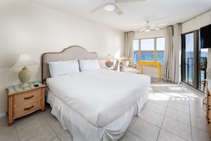 A hotel room with a large bed, a nightstand with a lamp, a chair, and a view of the ocean through large windows.
