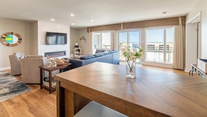 A modern living room with a wooden dining table, a blue sofa, a flat-screen TV, and a large window with a view of the beach.