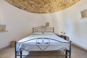 Ein Einzelbett mit Metallgestell und weißer Bettwäsche, ein kleiner Nachttisch mit Lampe sowie eine Steinwand oberhalb des Bettes.