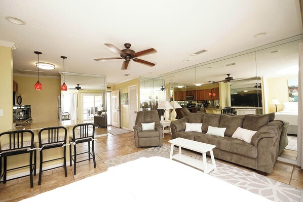 A spacious living room with a large gray sofa, a white coffee table, and a dining area with black chairs. A ceiling fan and pendant lights are visible. - Cozy Coastal Studio | Near Pool & Beach (Destin)