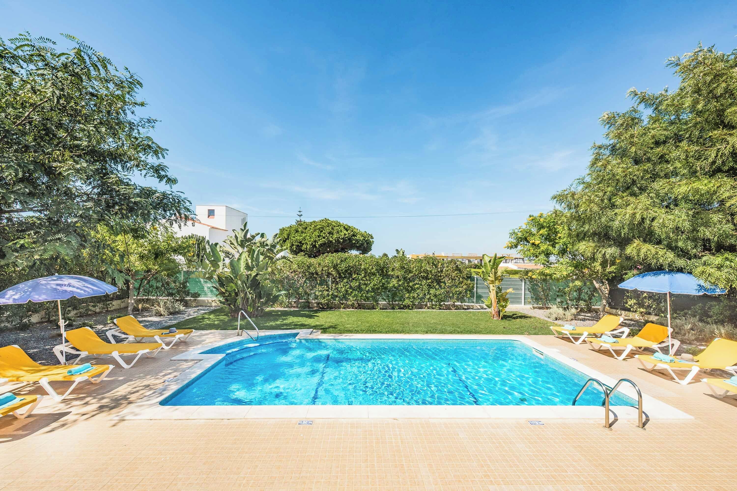 A swimming pool with clear blue water, surrounded by yellow lounge chairs and blue umbrellas.