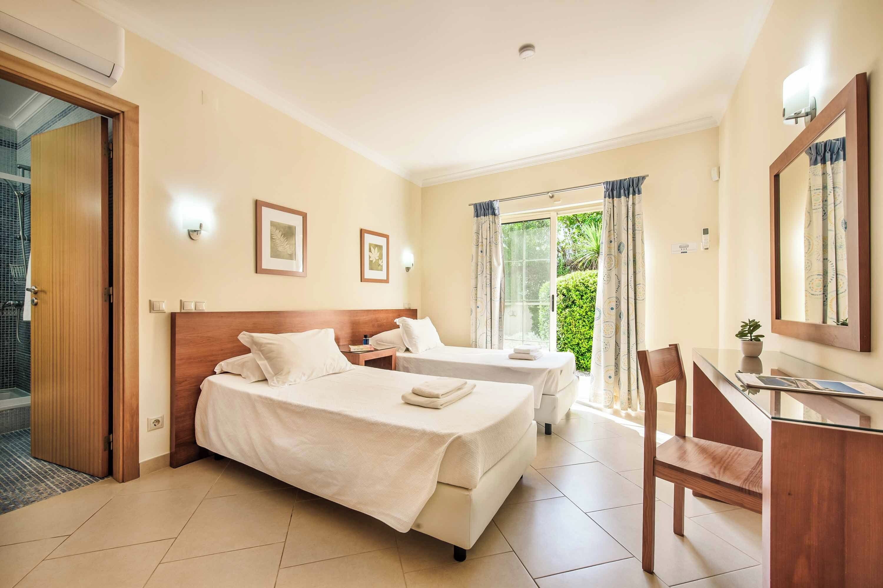 Habitación de hotel con cama, escritorio y silla. Se ve un ventanal con cortinas y vistas al exterior con zonas verdes.