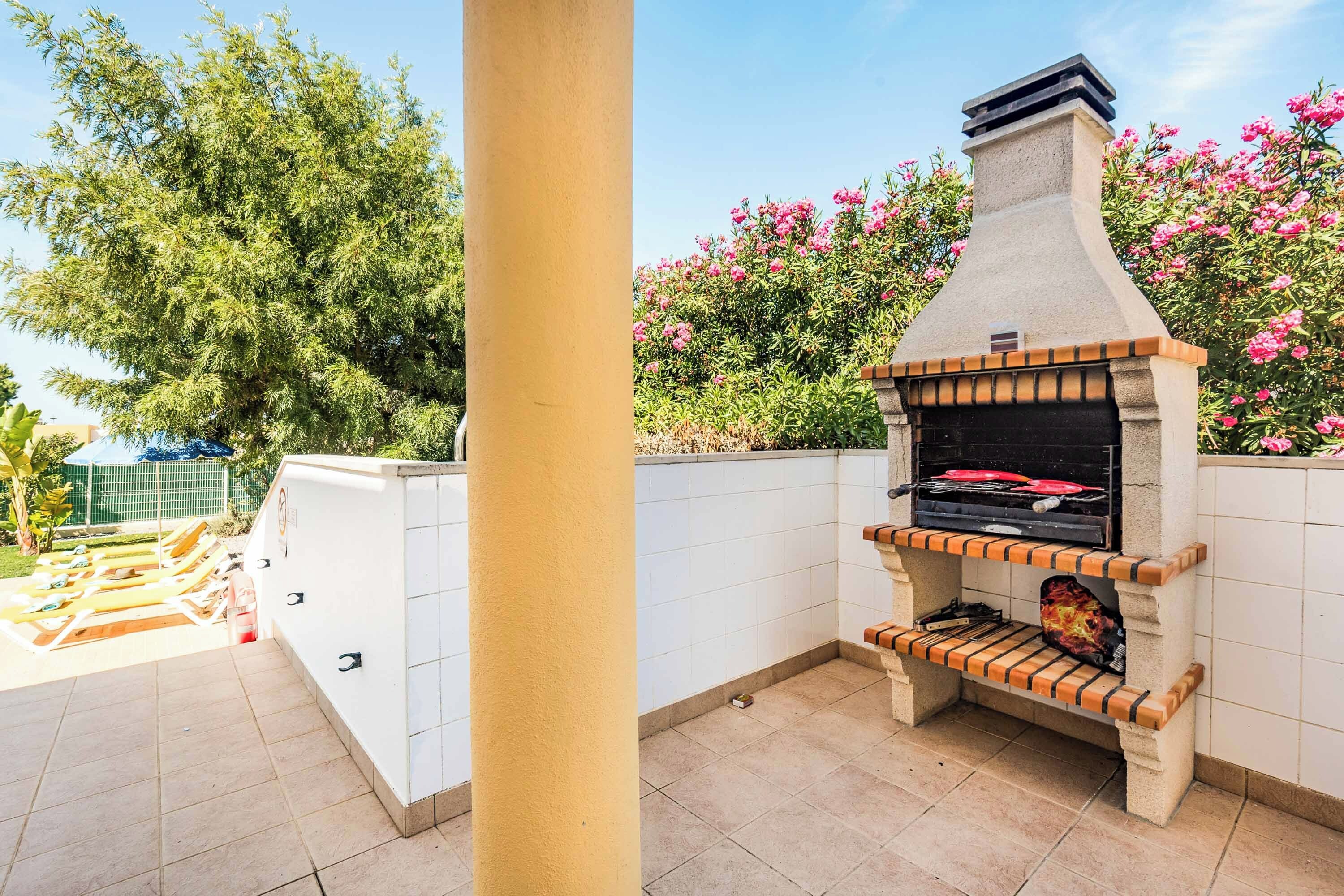 Un espace barbecue extérieur doté d’un four en pierre, d’une terrasse carrelée et d’une plateforme en bois avec des chaises longues.