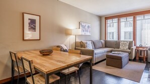 Un salon moderne avec une table à manger en bois, un canapé et une table basse. Des tableaux encadrés ornent le mur, et une fenêtre est équipée de stores.