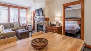 Une chambre d’hôtel dotée d’une cheminée en pierre, d’un téléviseur fixé au-dessus, d’un lit avec du linge de maison blanc, d’une table à manger en bois et d’une grande fenêtre avec des stores.
